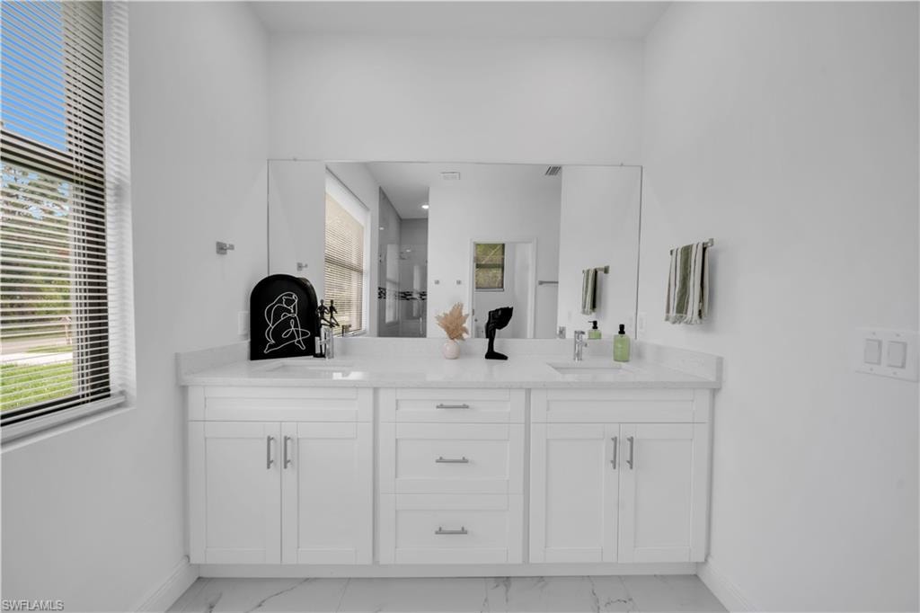 White cabinetry with brushed nickel hardware, expansive countertop with an integrated sink, large wall-mounted mirror, and a window with horizontal blinds