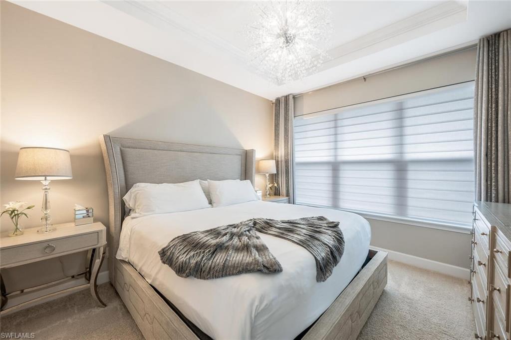 Tray ceiling with a decorative light fixture, large window with layered window treatments, upholstered headboard, textured carpeting, and painted walls