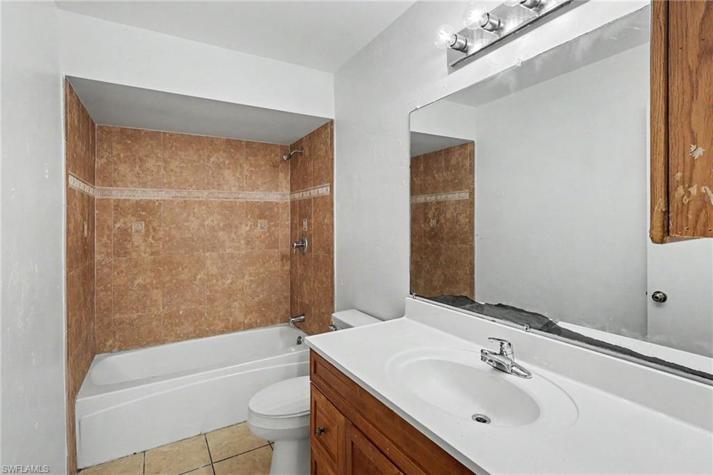 Bathroom featuring a tiled shower surround with decorative accent band, a white bathtub, a toilet, a wood-finish vanity with an integrated sink and chrome faucet, and a large wall-mounted mirror with a vanity light fixture