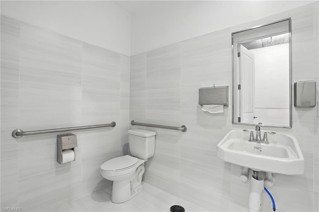 Restroom featuring bright white wall and floor tiles, a wall-mounted sink with chrome faucet, a white toilet, and stainless steel grab bars