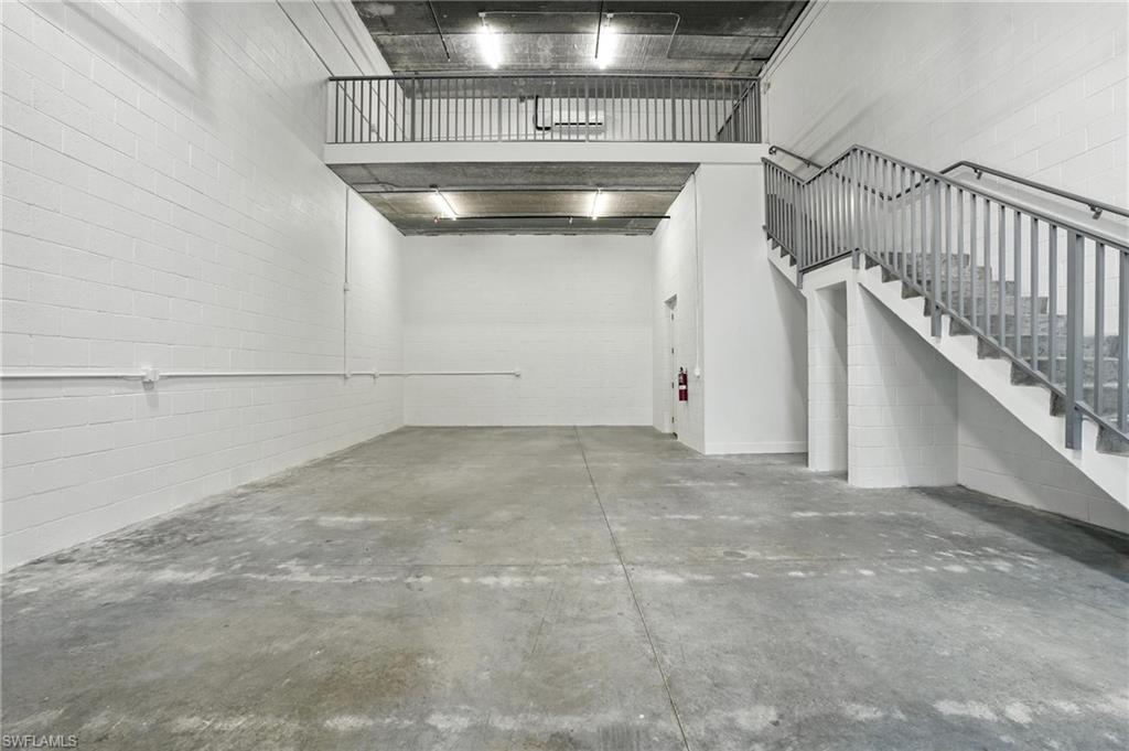Spacious interior featuring a concrete floor, painted concrete block walls, exposed concrete ceiling, and a prominent staircase with metal railings leading to an upper mezzanine