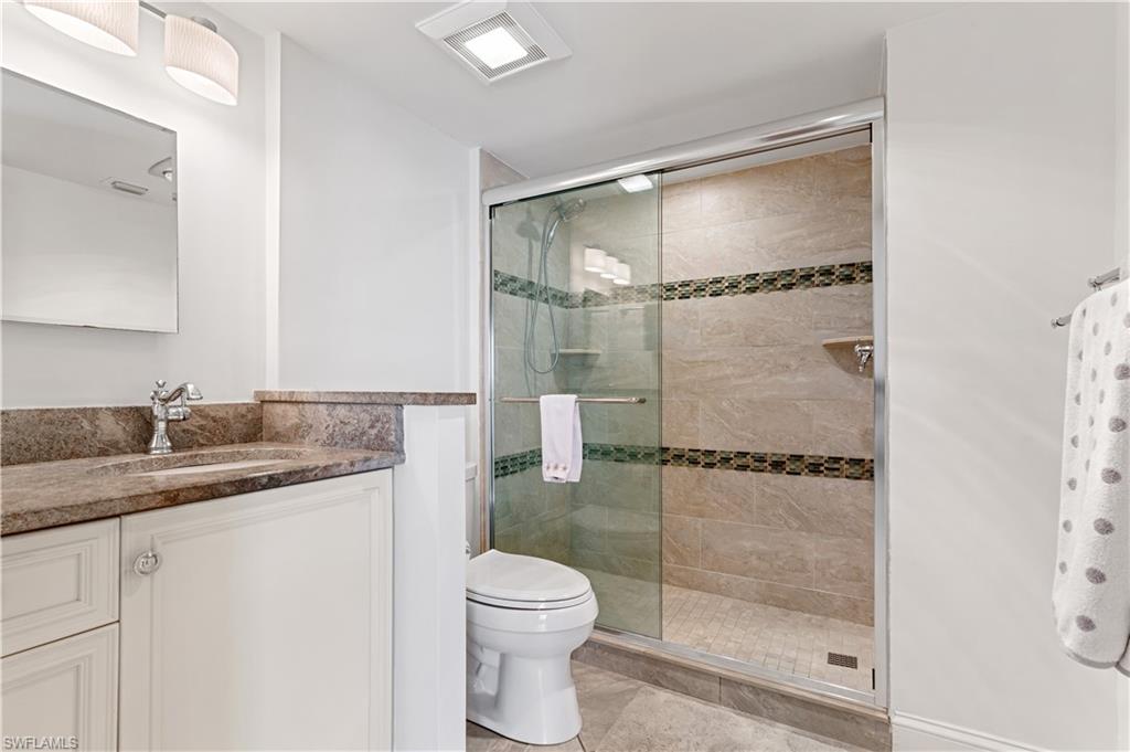 Bathroom featuring a glass-enclosed shower with decorative tile accents, a single vanity with a stone countertop and integrated sink, a commode, and overhead lighting