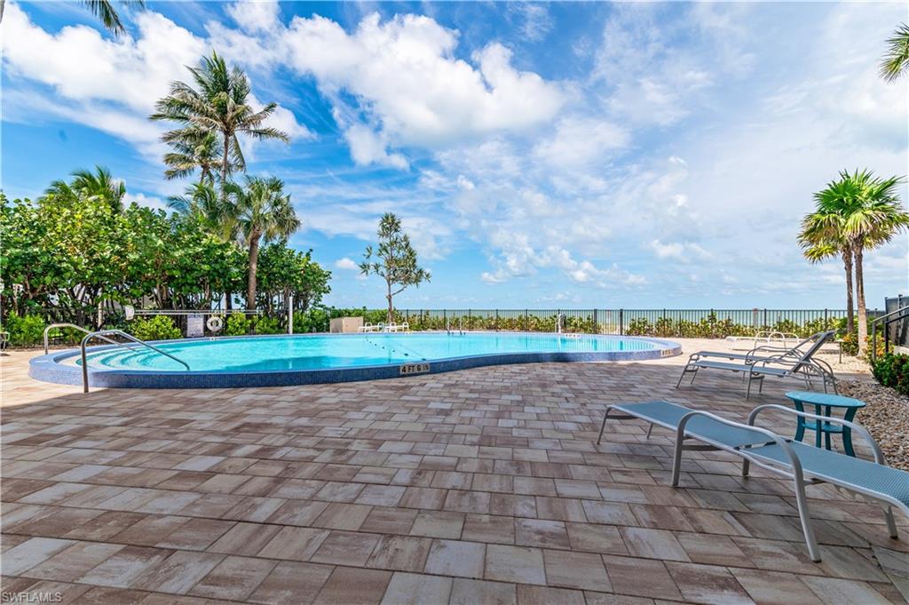 Curved swimming pool with blue mosaic tile border, surrounded by extensive paver decking, lush tropical landscaping, and a dark metal fence