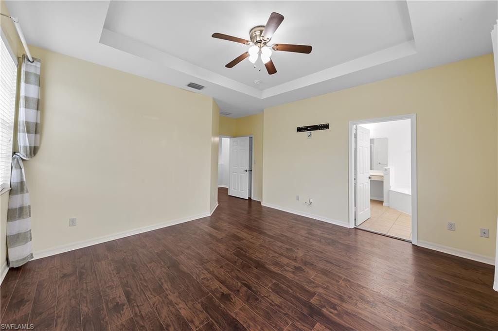 Unfurnished bedroom featuring dark wood-type flooring, a ceiling fan, a tray ceiling, and ensuite bath
