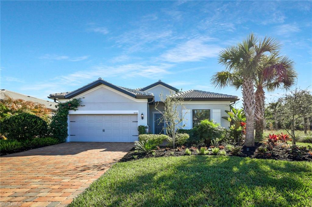 Modern stucco exterior with gray tile roof, decorative garage door, paver driveway, and lush tropical landscaping