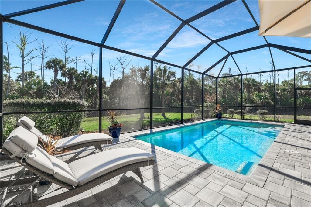 Screened pool enclosure featuring a rectangular swimming pool with steps, paver decking, and a backdrop of mature trees