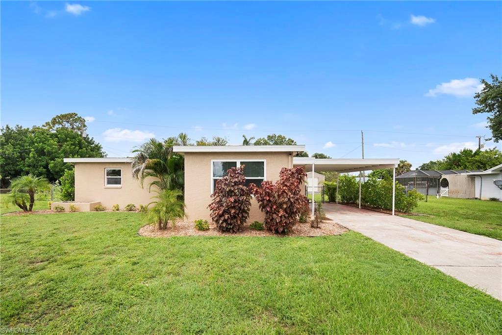 Single-story residence featuring a painted block exterior, manicured front lawn, established tropical landscaping, covered carport, and concrete driveway