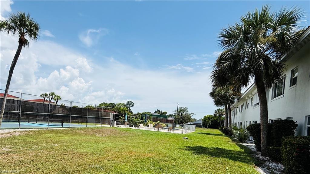 Expansive green space featuring a tennis court with chain-link fencing, mature palm trees, and a low-rise building with multiple windows