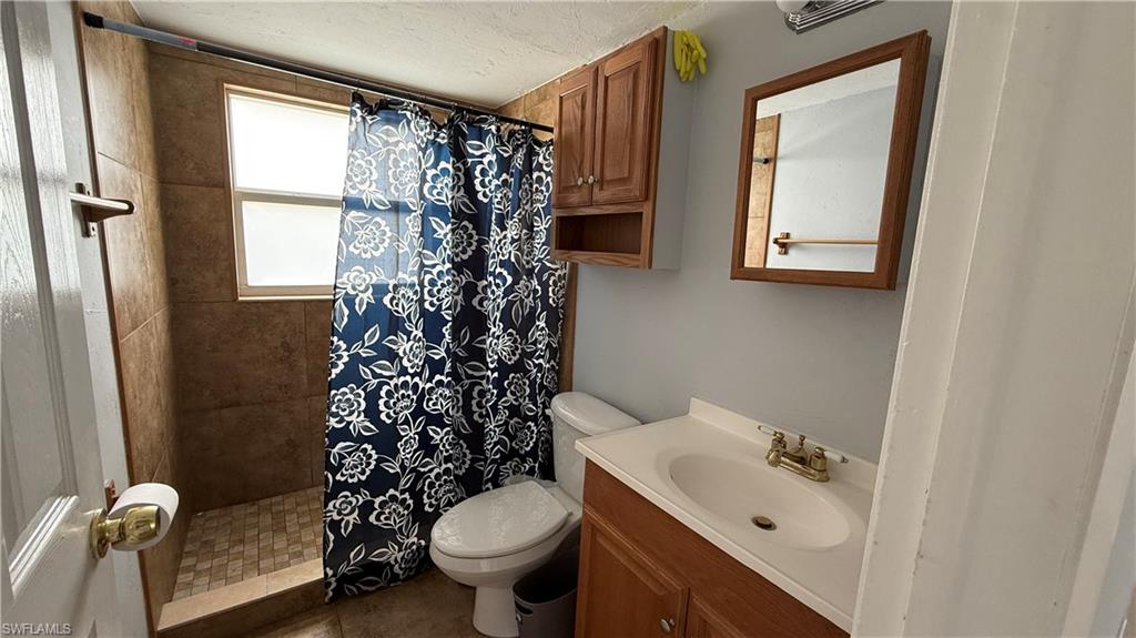 Full bathroom featuring ceramic tile flooring, wood-finish vanity with white porcelain sink, brass faucet hardware, toilet, and walk-in shower with tile surround and decorative shower curtain rod