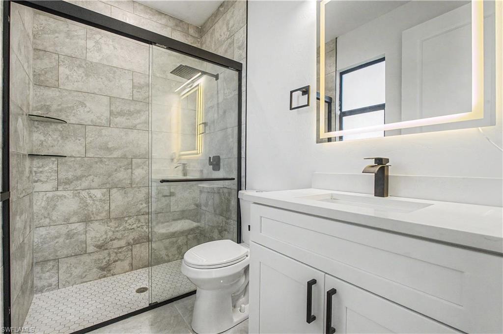 Bathroom featuring a walk-in shower with large format tile walls and a hexagonal tile floor, a white vanity with a dark bronze faucet, and an illuminated mirror