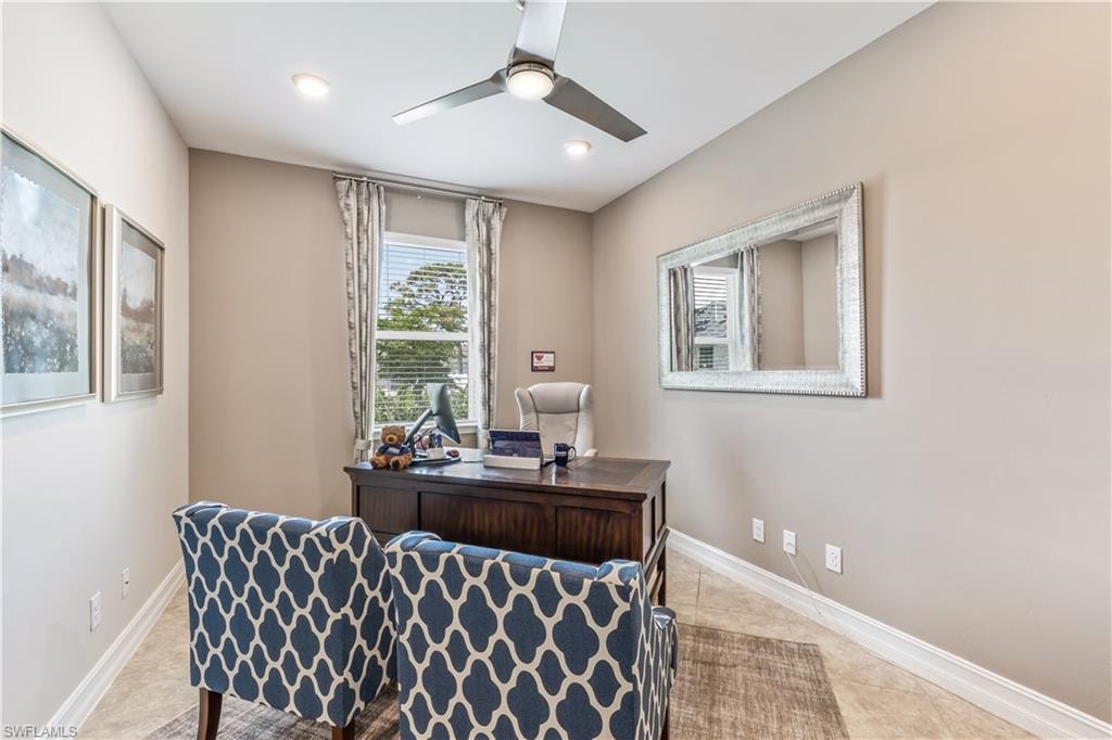 Dedicated home office featuring a wood-finish desk with ample storage, patterned upholstered chairs, large mirror, recessed lighting, ceiling fan, and natural light from a double-hung window with custom drapery