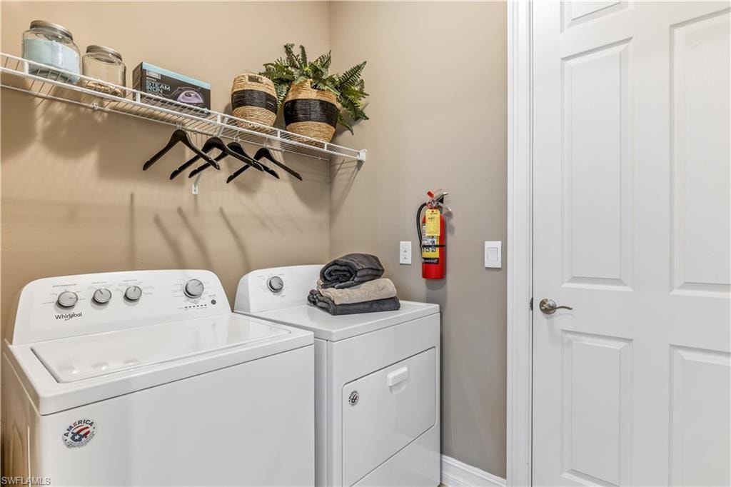Dedicated laundry room featuring side-by-side washer and dryer with white finish, stainless steel countertop workspace, wall-mounted shelving with storage baskets, black wall hooks for hanging supplies, fire extinguisher, and neutral painted walls with direct access via six-panel interior door