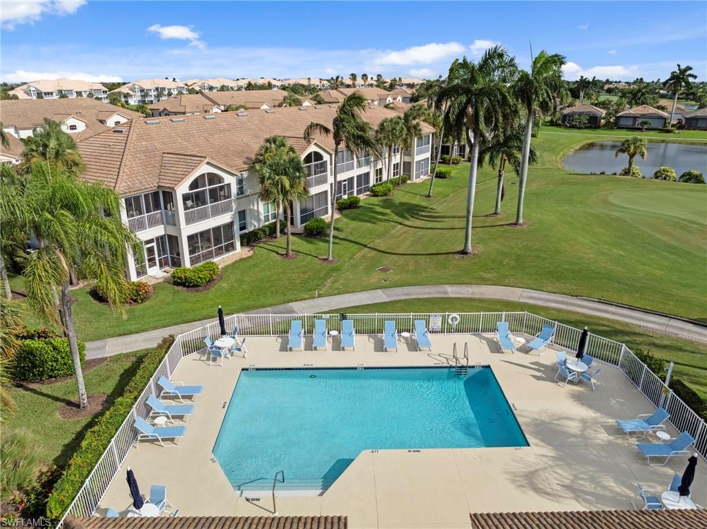 Resort-style community pool and deck featuring a sparkling blue swimming pool, lounge seating with umbrellas, mature palm trees, manicured landscaping, and multi-story residential buildings with two-story architecture overlooking the expansive grounds and adjacent waterfront