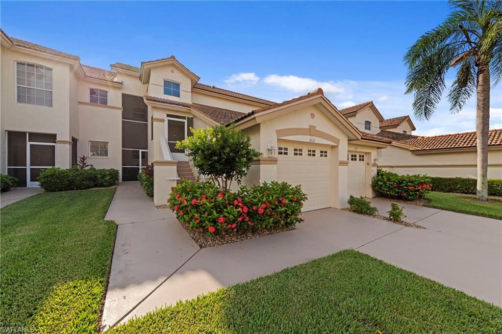 Charming Mediterranean-style residence featuring a pristine manicured lawn, mature landscaping with flowering shrubs, and a spacious concrete driveway leading to a two-car garage with decorative hardware