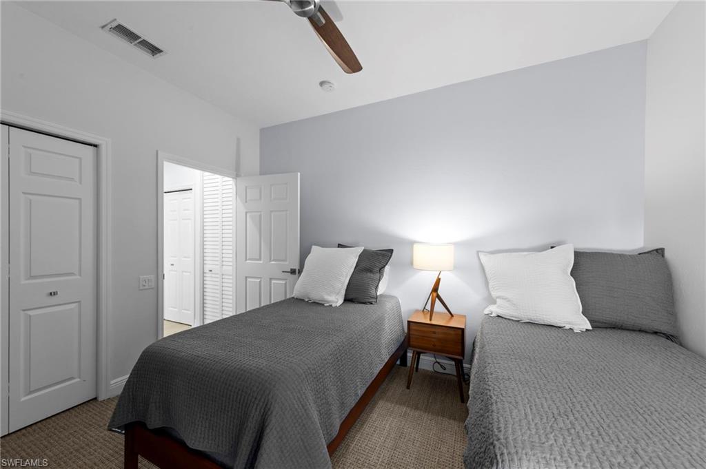 Comfortable guest bedroom featuring two twin beds with charcoal upholstered headboards and white bedding, wood-finish nightstand with integrated lighting, wood-finish flooring, ceiling fan, and direct access to an ensuite bathroom