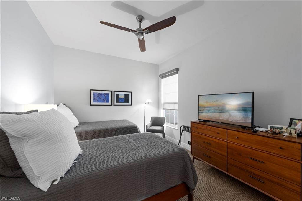 Spacious bedroom featuring dual beds with coordinating gray bedding, wood-finish dresser with integrated television mount, ceiling fan with dark blades, carpeted flooring, and abundant natural light from sliding glass doors