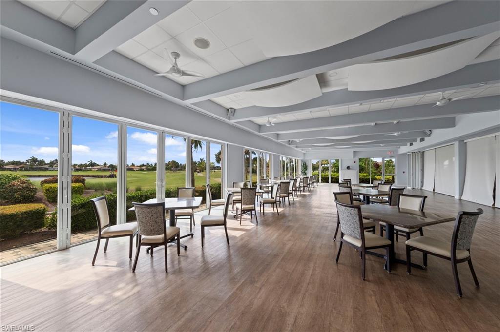Expansive open-concept dining and lounge area featuring floor-to-ceiling glass doors and windows showcasing pastoral grounds, wide-plank wood-finish flooring throughout, coffered ceiling with recessed lighting, and contemporary upholstered seating arrangements positioned to maximize natural light and sightlines