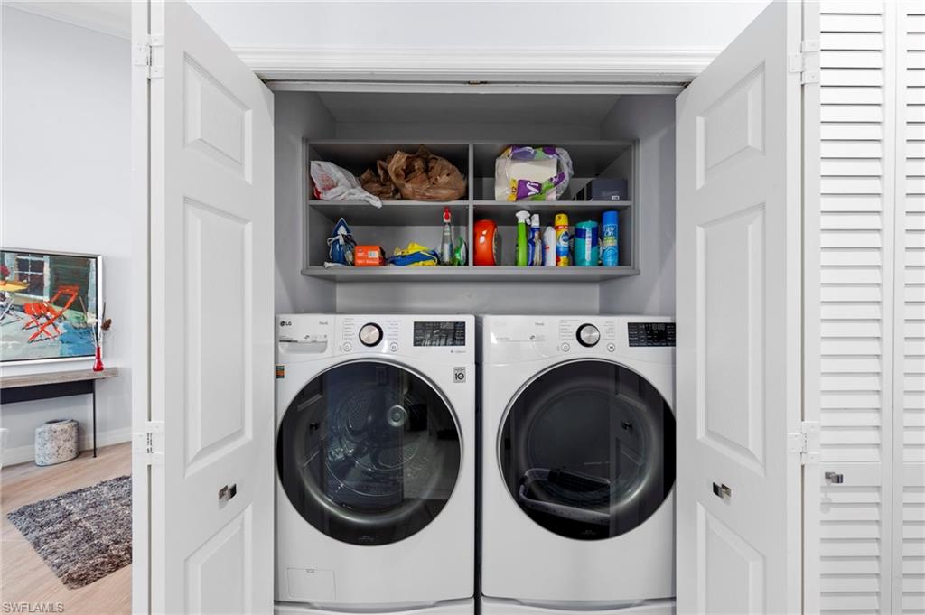Dedicated laundry closet featuring side-by-side front-load washer and dryer with modern stainless steel finishes, overhead shelving with ample storage for linens and cleaning supplies, and louvered closet doors for space-efficient access