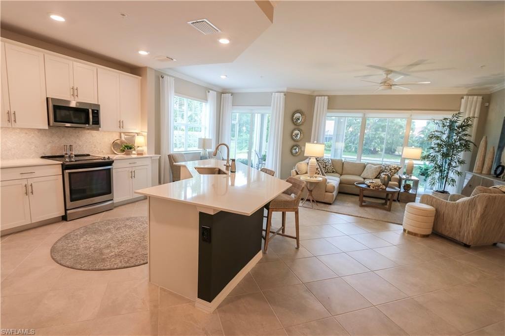 Open-concept living area featuring light-toned tile flooring, white cabinetry, stainless steel appliances, a large kitchen island with counter seating, and expansive windows