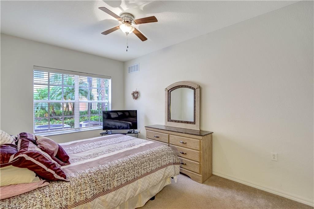 Spacious bedroom with expansive windows flooding the space with natural light, ceiling fan with integrated lighting, wood-finish dresser with attached mirror, and neutral carpeting throughout