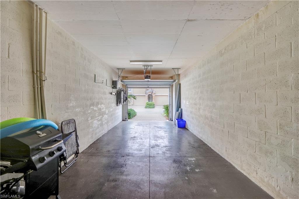 Spacious garage featuring concrete flooring, white brick walls, overhead storage systems, and a roll-up door with natural light access to the driveway