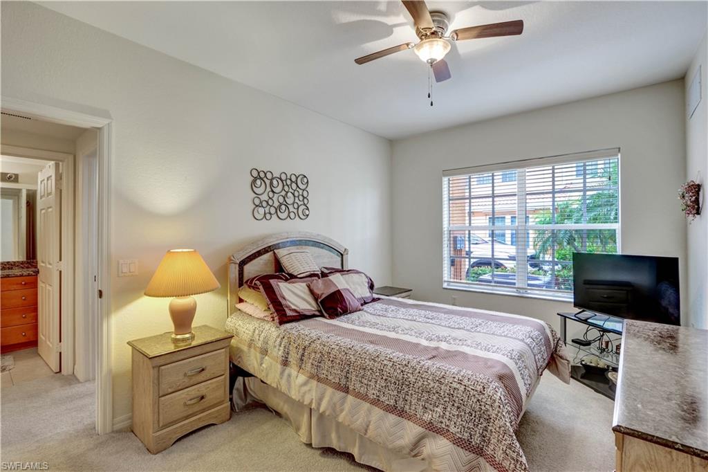 Bright secondary bedroom featuring a wood-finish nightstand with integrated lighting, carpet flooring, ceiling fan with integrated light fixture, and large multi-pane window with plantation shutters allowing abundant natural light