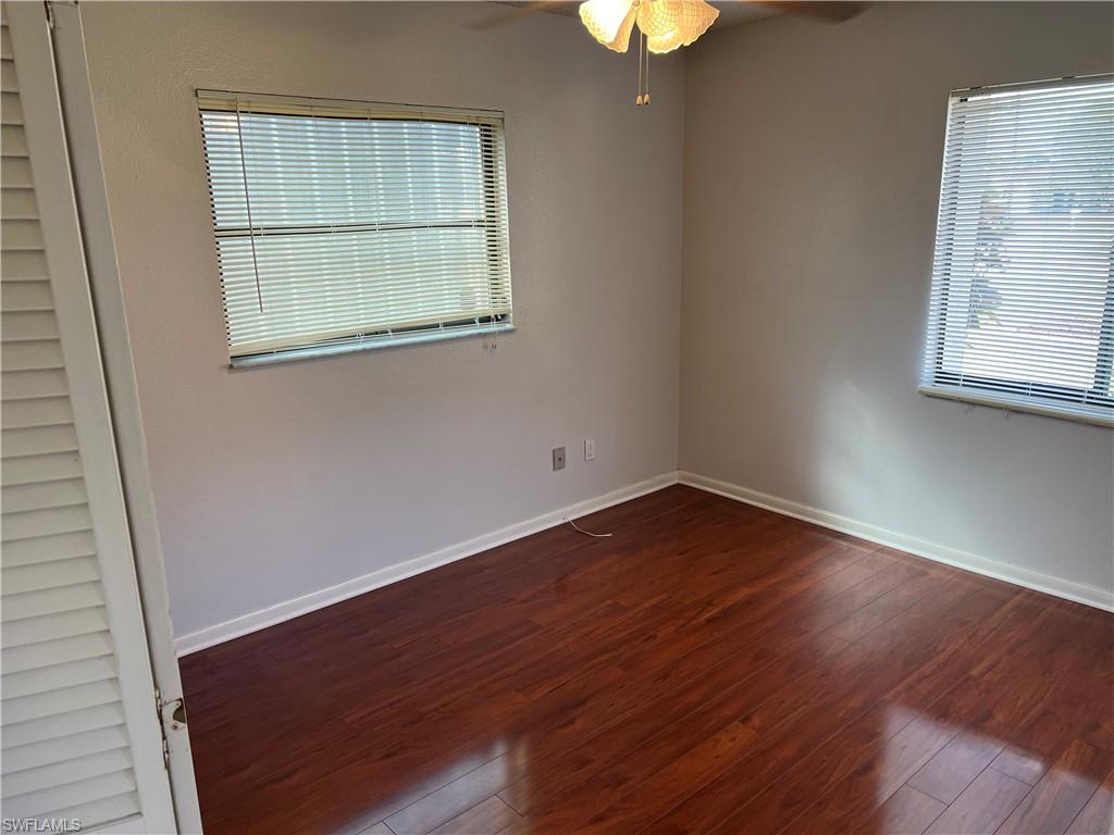 Bright secondary bedroom featuring rich wood-finish flooring, neutral wall finishes, and abundant natural light from two large windows with horizontal blinds