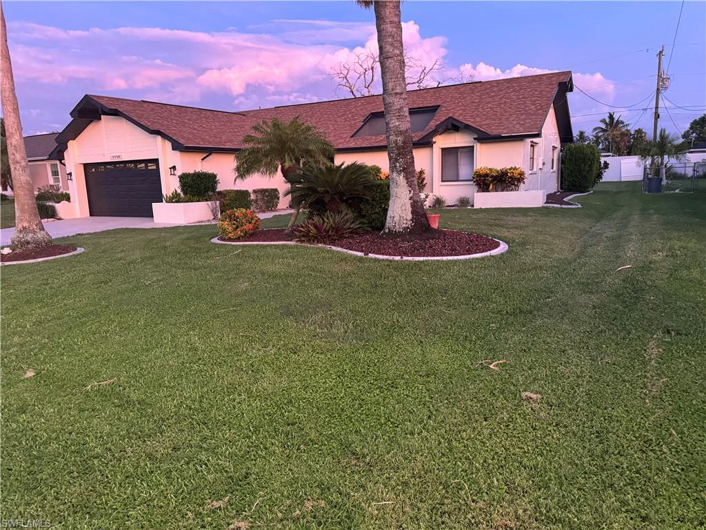 Charming single-story home featuring a sprawling manicured lawn, mature landscaping with curved planting beds, attached two-car garage with dark finish, and a pink stucco exterior