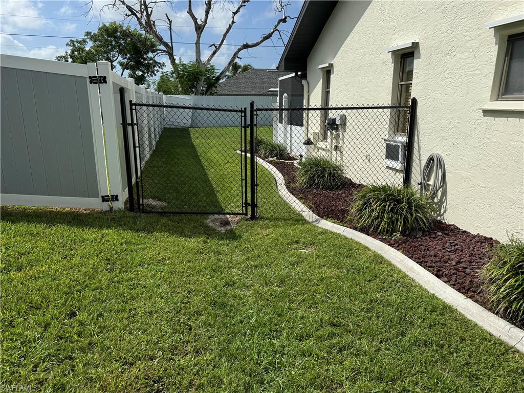Fenced backyard oasis featuring a lush, level lawn with mature landscaping, black chain-link gate entry, decorative white edging, and mulched planting beds with ornamental grasses