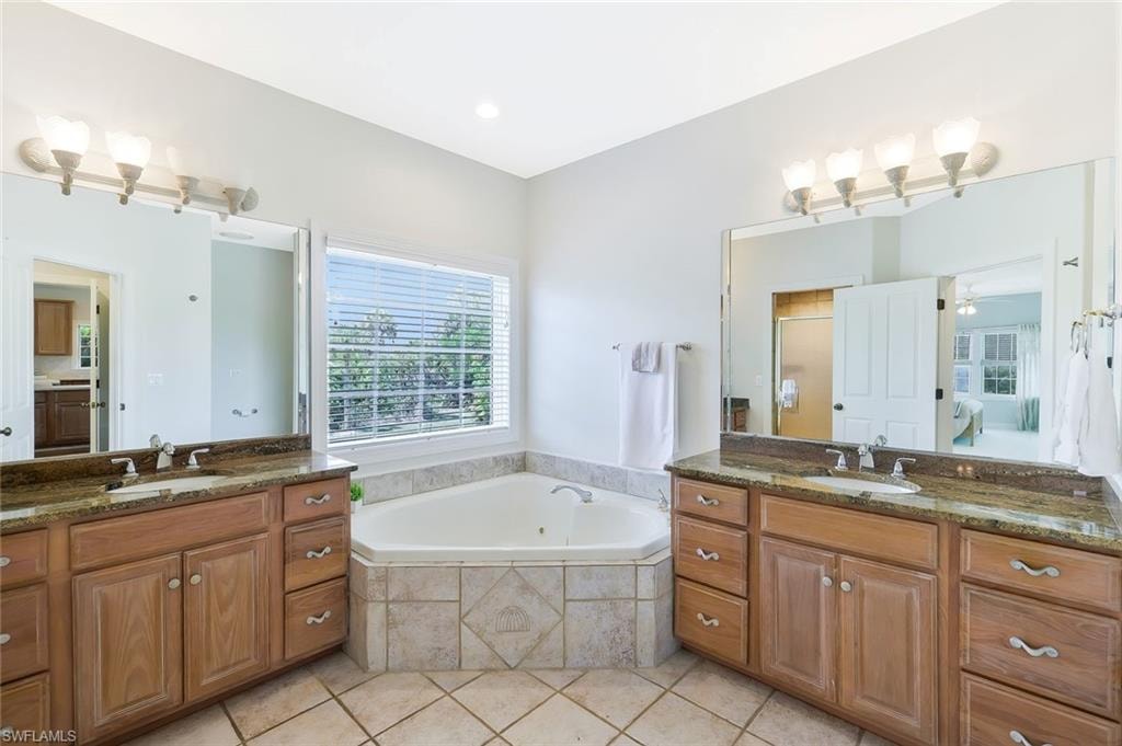 Spa-inspired primary bathroom featuring dual vanities with wood cabinetry and granite countertops, corner soaking tub with tile surround, dual illuminated mirrors, and abundant natural light from large window