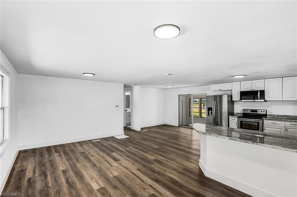 Open-concept living area with wood-finish flooring, recessed lighting, white walls, and a kitchen featuring white cabinetry, stainless steel appliances, and a granite-style countertop peninsula