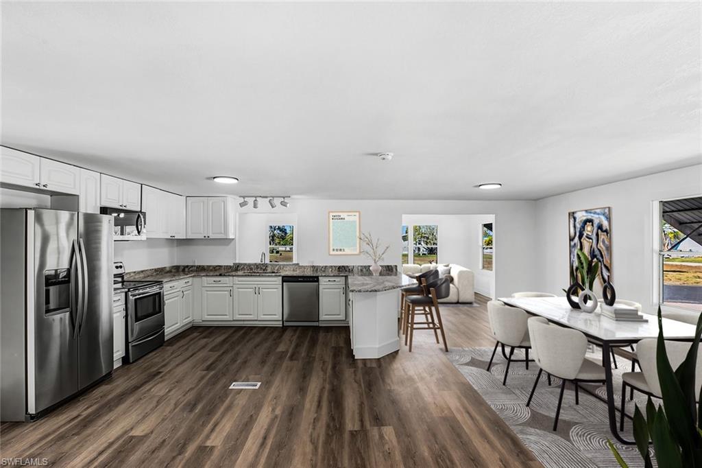 Open-concept kitchen and dining area featuring stainless steel appliances, white cabinetry with black countertops, a large island with seating, wood-finish flooring throughout, recessed lighting, and abundant natural light from multiple windows and glass doors