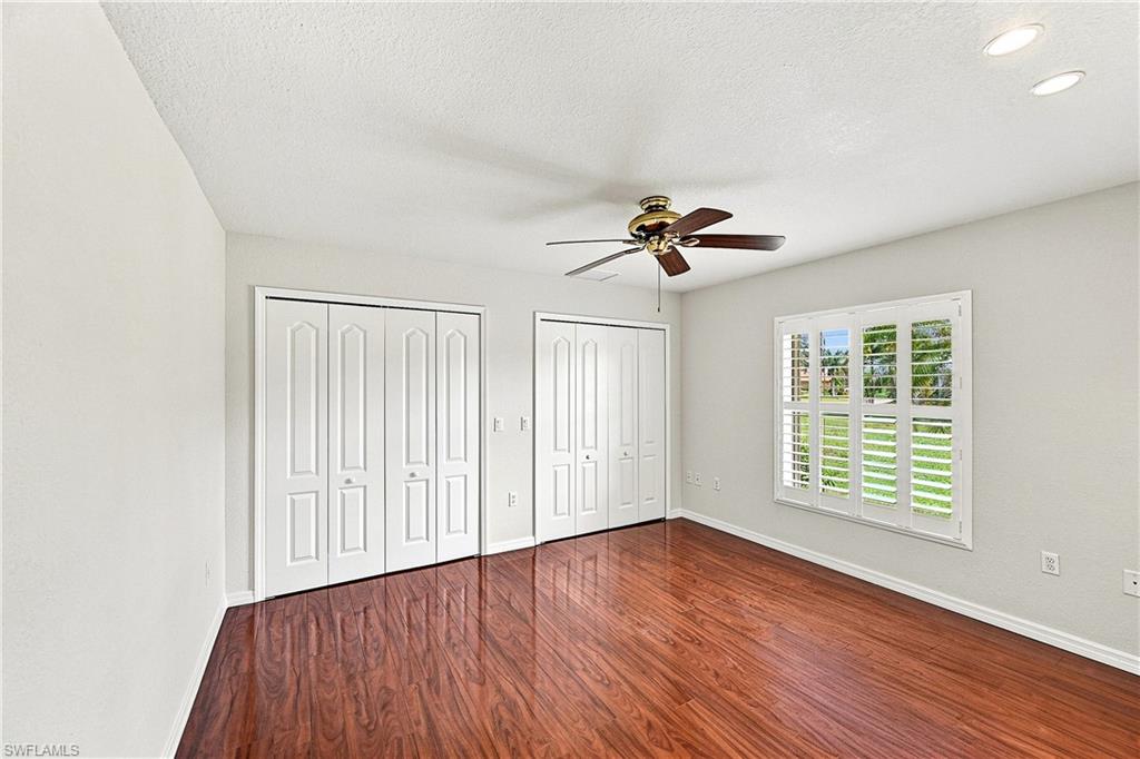 Spacious guest bedroom showcasing rich wood-finish flooring, white panel closet doors with multiple storage sections, white plantation shutters on expansive windows, ceiling fan, and bright recessed lighting