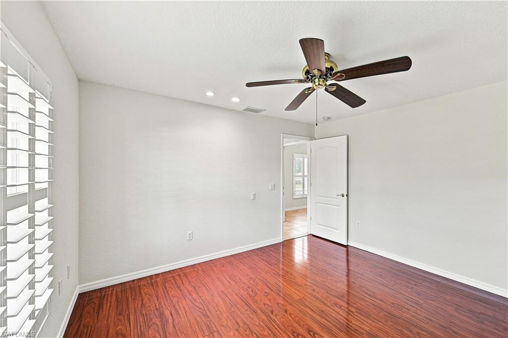 Spacious bedroom featuring rich wood-finish flooring, recessed ceiling lighting, ceiling fan with brushed metal and dark blade finish, and abundant natural light from shuttered windows