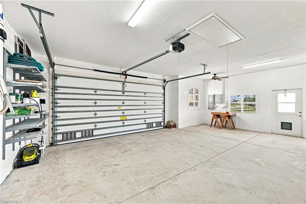 Spacious garage featuring a hurricane rated garage door, concrete flooring, fluorescent lighting, and built-in shelving, pull-down stairs to attic area. Windowed corner area with fan is workshop space, with electric panels pre-wired for portable generator.