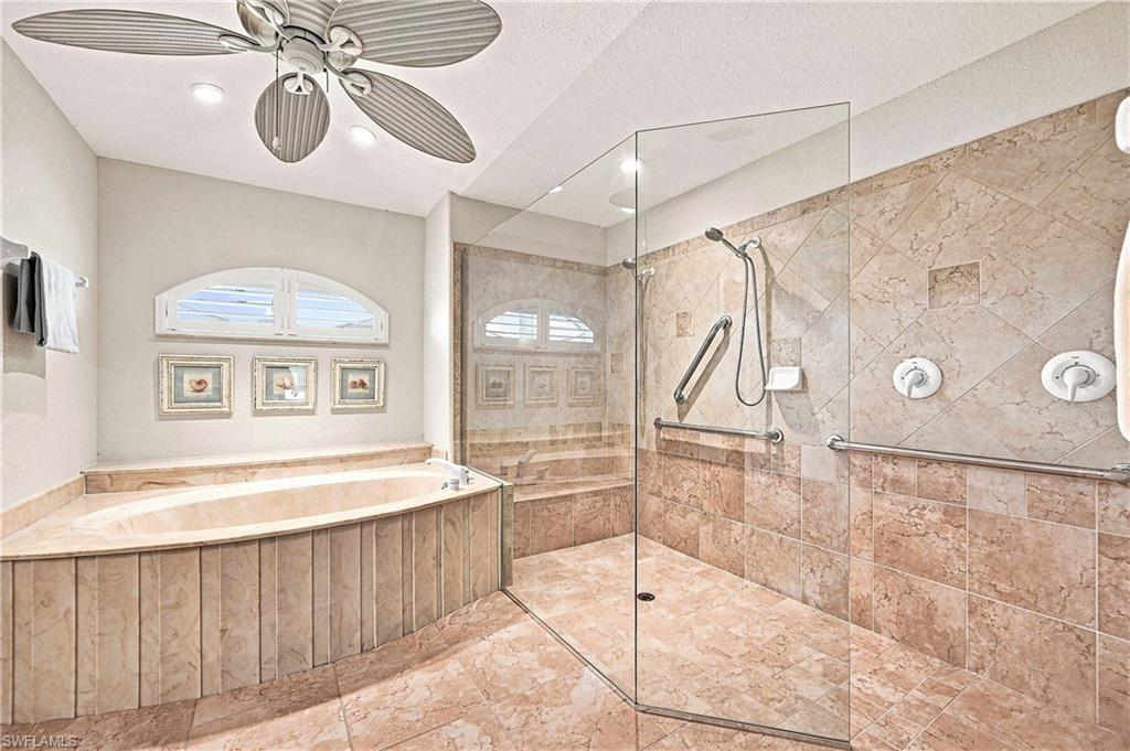 Full-tile primary bathroom featuring a built-in soaking tub, walk-in shower with glass enclosure, arched window with plantation shutters, and ceiling fan with integrated lighting