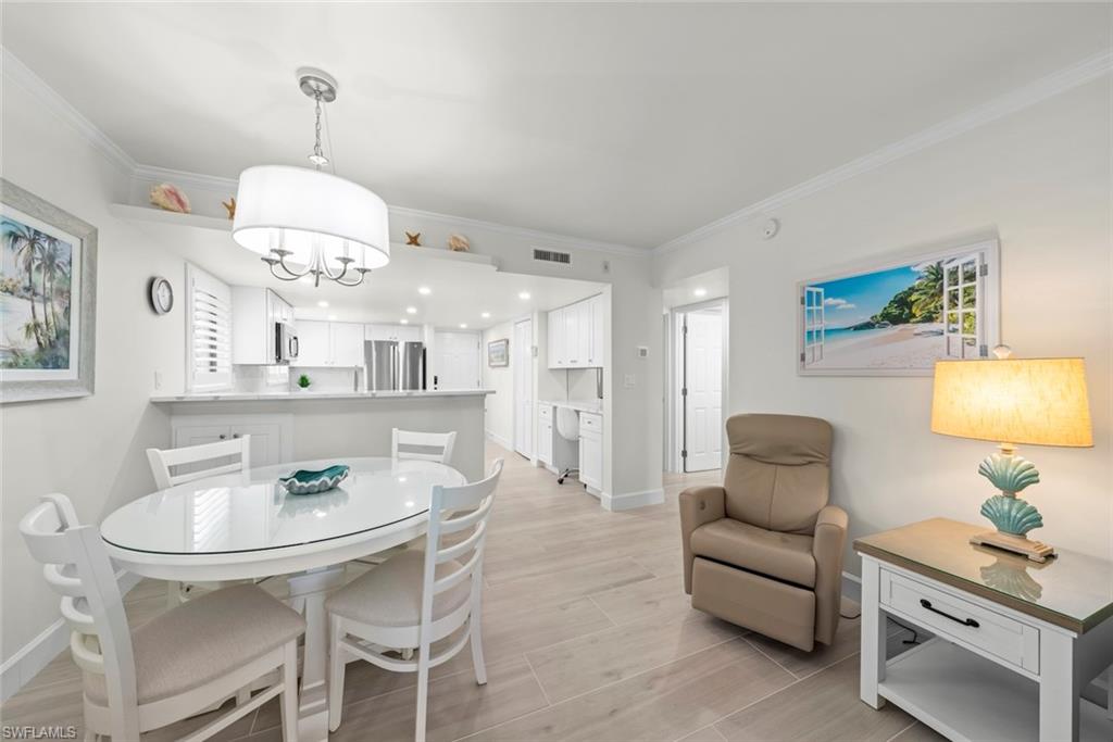 Open-concept living area featuring light-toned flooring, a white dining table with four chairs, a kitchen with white cabinetry and stainless steel appliances, a breakfast bar, and a contemporary pendant light fixture