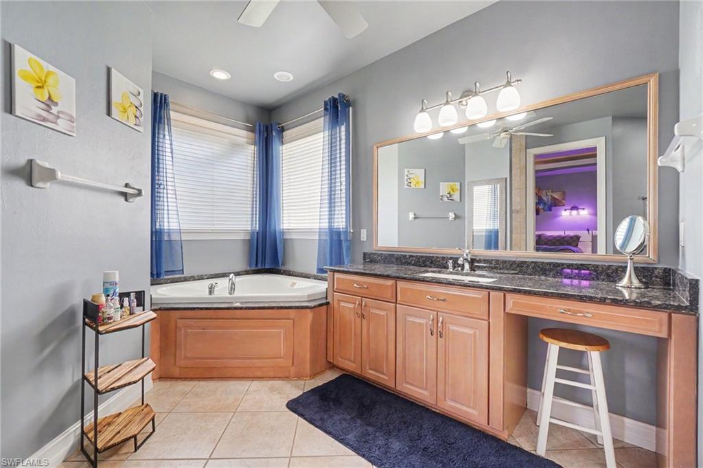 Bathroom featuring a corner soaking tub, dual windows with blinds, wood-finish vanity with an integrated sink, dark granite-style countertop, and a large wall-mounted mirror with overhead lighting
