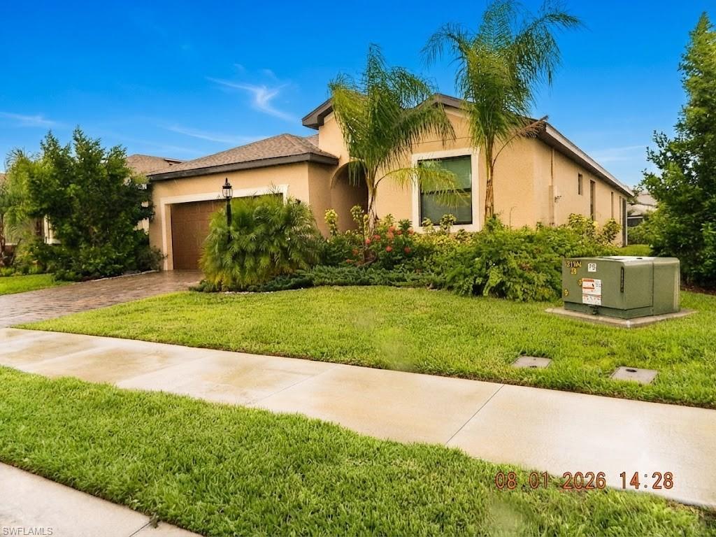 Stucco-finished residence with barrel tile roof accents, featuring a two-car garage, mature landscaping with tropical palms and established shrubs, manicured lawn, and concrete driveway with adjoining sidewalk