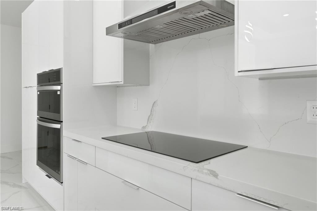 Integrated stainless steel double ovens, sleek induction cooktop, modern white cabinetry, stainless steel range hood, and continuous countertop and backsplash material