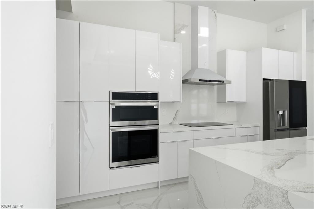 Contemporary kitchen featuring white flat-panel cabinetry, a built-in double oven, stainless steel range hood, and a spacious island with a stone countertop
