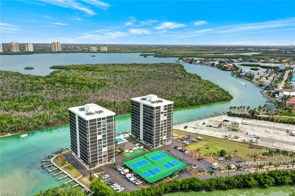 Two high-rise structures featuring multiple balconies, surrounded by water with boat docks, two tennis courts, and a parking area, all set against a backdrop of natural waterways and extensive greenery