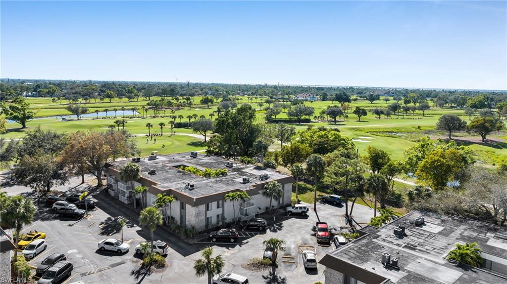 Aerial perspective of a multi-story building featuring an asphalt parking lot, mature palm trees, and an expansive golf course backdrop with water features
