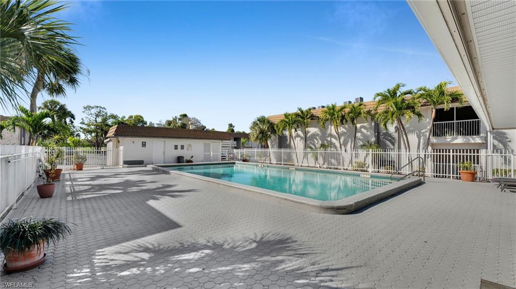 Resort-style community pool and deck featuring crystal-clear turquoise waters, expansive stamped concrete decking with defined shade patterns, mature palm tree landscaping, and a dedicated pool house structure