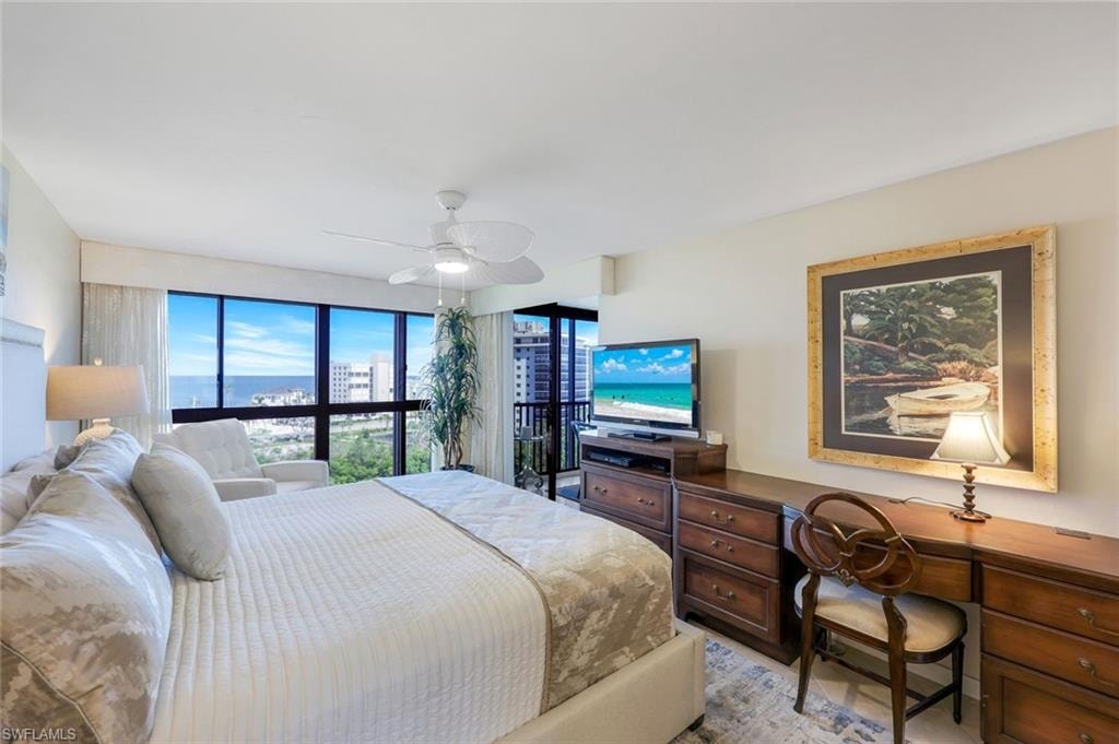 Spacious bedroom suite featuring floor-to-ceiling sliding glass doors opening to a private balcony with ocean vistas, a ceiling fan with integrated lighting, wood-finish dresser with matching desk, framed artwork, and striped bedding on a generous bed frame