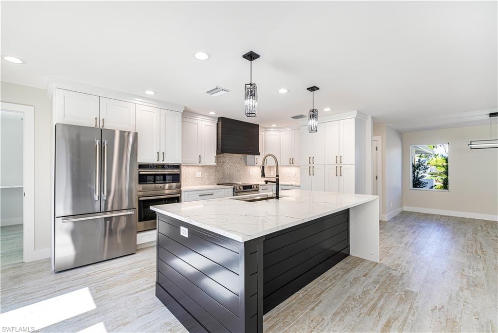 Spacious kitchen featuring a central island with a waterfall-edge countertop and integrated sink, stainless steel appliances including a French door refrigerator and built-in oven, white shaker cabinetry with ample storage, recessed lighting, and wood-finish flooring