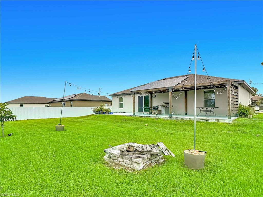 Expansive green lawn with a stone-surround fire pit, featuring a covered patio with string lighting, multiple solar panels on the roof, and a white privacy fence
