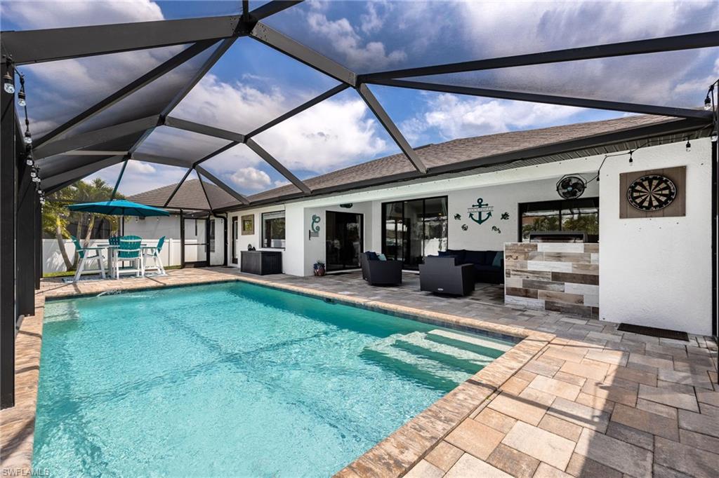 Screened pool enclosure featuring a rectangular pool with integrated steps, paver decking, a built-in outdoor kitchen with stone veneer, and a covered patio area