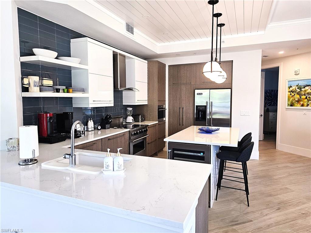 Kitchen with pendant lighting, open shelves, white cabinetry, wall chimney range hood, and a kitchen bar