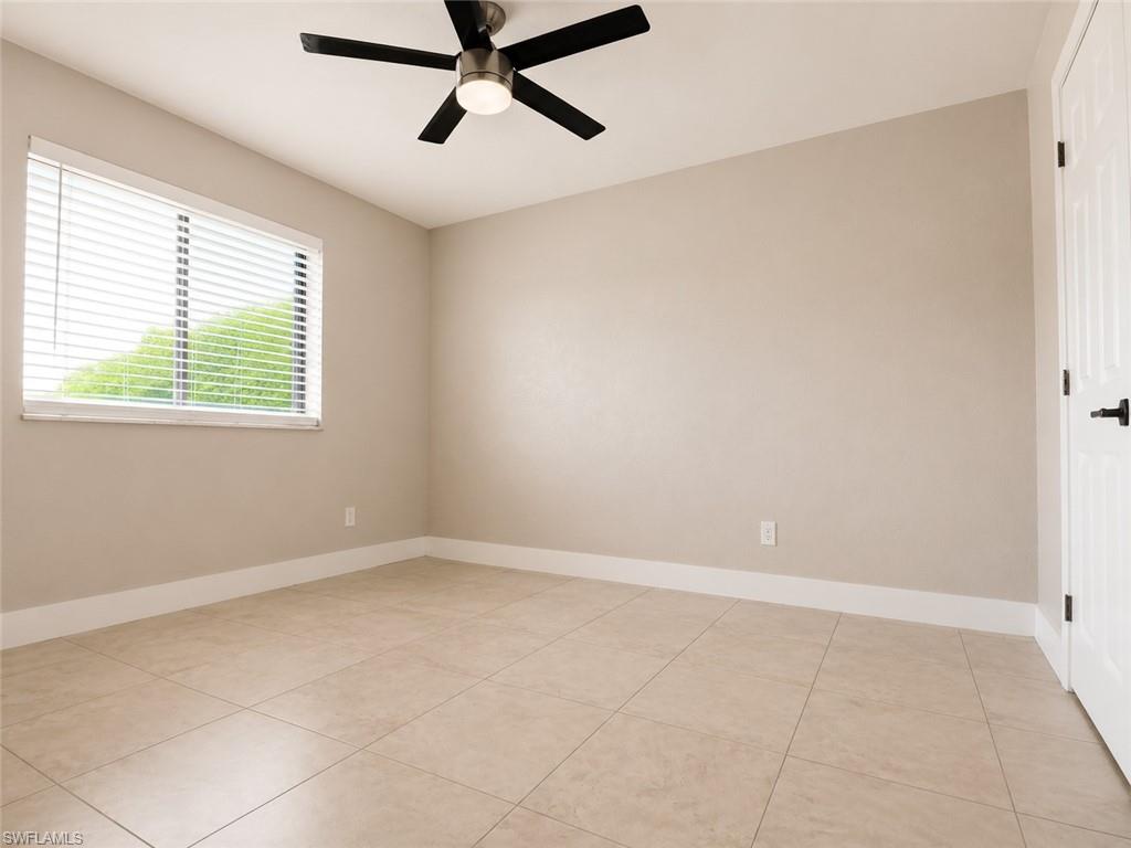 Bright, open room featuring neutral taupe walls, tile flooring throughout, large window with plantation shutters providing natural light, ceiling fan with integrated lighting, and white trim detailing